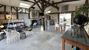 Amplia sala de estar con vigas de madera a la vista, chimenea de piedra, comedor con mesa de vidrio y un rincón de descanso acogedor con una silla colgante y cojines.