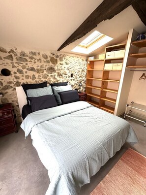 A bedroom with a stone wall, a bed with a light blue bedspread, a wooden headboard, and a skylight above the bed. - Lou Sarrat - MIMBASTE (MIMBASTE)