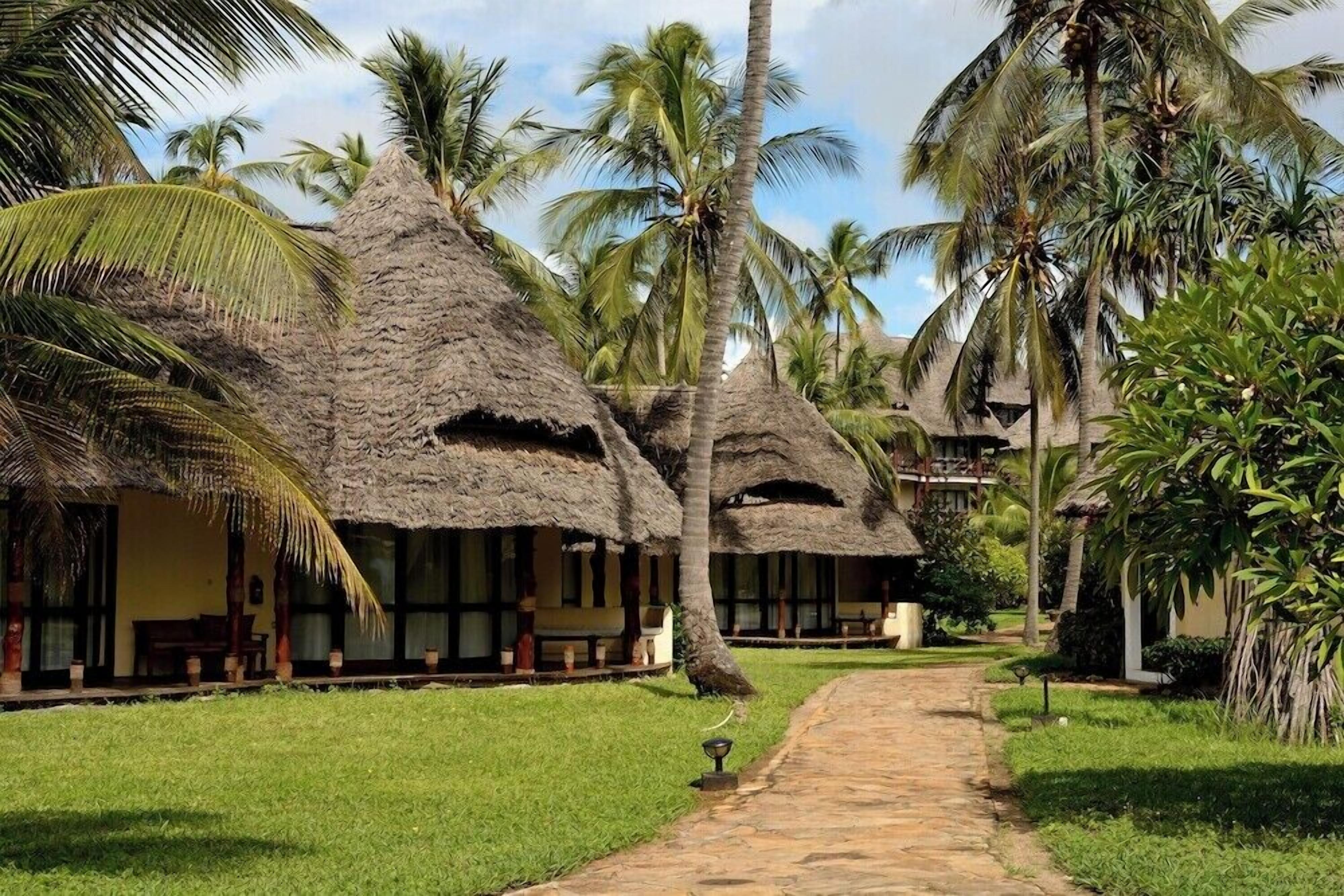 Uma trilha que leva a construções com telhados de palha, palmeiras e vegetação.