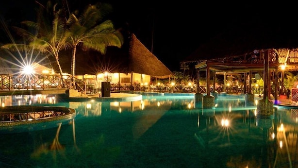 A nighttime resort pool area with a thatched-roof structure, palm trees, and illuminated pool lights. - Ocean Paradise Resort Zanzibar (Kiwengwa)