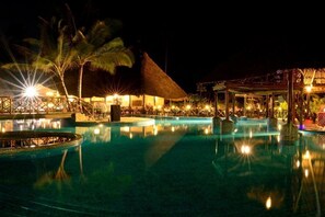 A nighttime resort pool area with a thatched-roof structure, palm trees, and illuminated pool lights. - Ocean Paradise Resort Zanzibar (Kiwengwa)