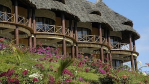 A building with multiple thatched-roof structures, a circular balcony, and lush gardens with various flowers. - Ocean Paradise Resort Zanzibar (Kiwengwa)