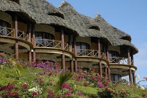 Un edificio con varias construcciones de techo de paja, un balcón circular y exuberantes jardines con diversas flores.