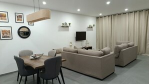 A modern living room with a sofa set, a dining table with chairs, a flat-screen TV, and wall-mounted shelves with decorative items. - Exclusive location, close to the Mall and main attraction. (San Salvador)