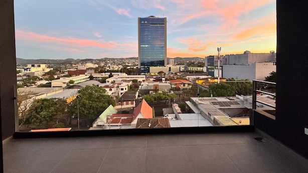 A panoramic view of a cityscape with a prominent skyscraper, various buildings, and a sunset sky. - Exclusive location, close to the Mall and main attraction. (San Salvador)