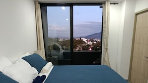 A hotel room with a large bed, a view of a cityscape through a sliding door, and a wooden wardrobe. - Exclusive location, close to the Mall and main attraction. (San Salvador)