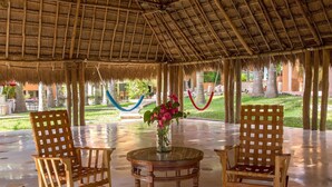 A covered outdoor seating area with two wooden rocking chairs and a glass table, surrounded by a thatched roof structure. - Hacienda Uxmal (Santa Elena)