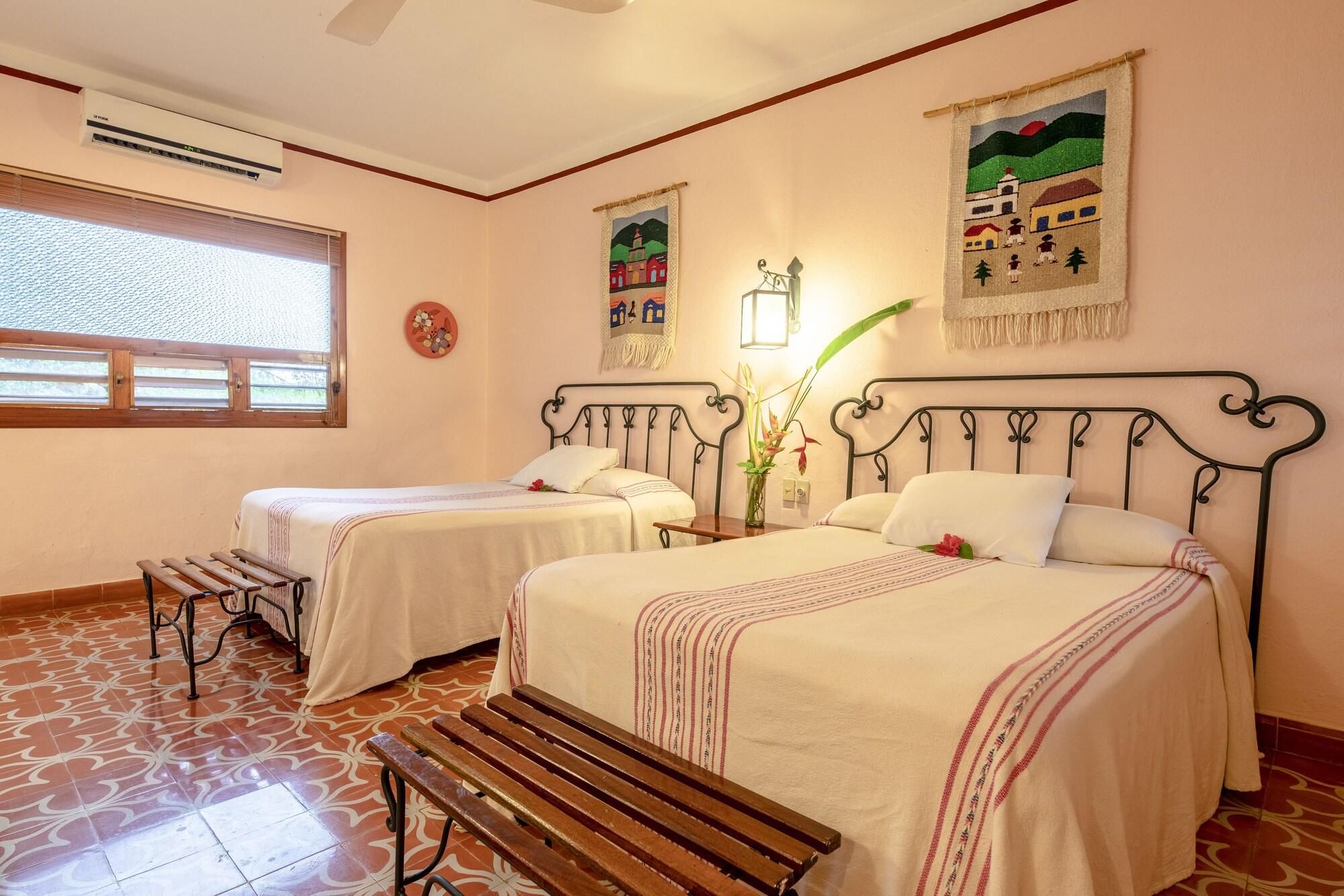 Una habitación de hotel con dos camas individuales, un banco, una ventana con persianas y obras de arte en las paredes.