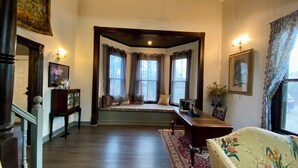 Una habitación con un amplio ventanal, suelo de madera, un banco con cojines, una mesita con un jarrón y una silla estampada con motivos florales.