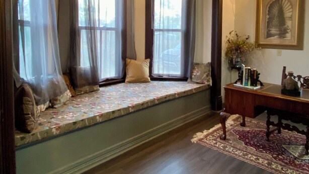 Una habitación con una zona de estar junto a un ventanal, un banco de madera, una alfombra y un cuadro en la pared.