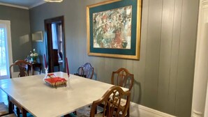 Comedor con una mesa blanca, sillas de madera, una lámpara de araña y un cuadro enmarcado en la pared.