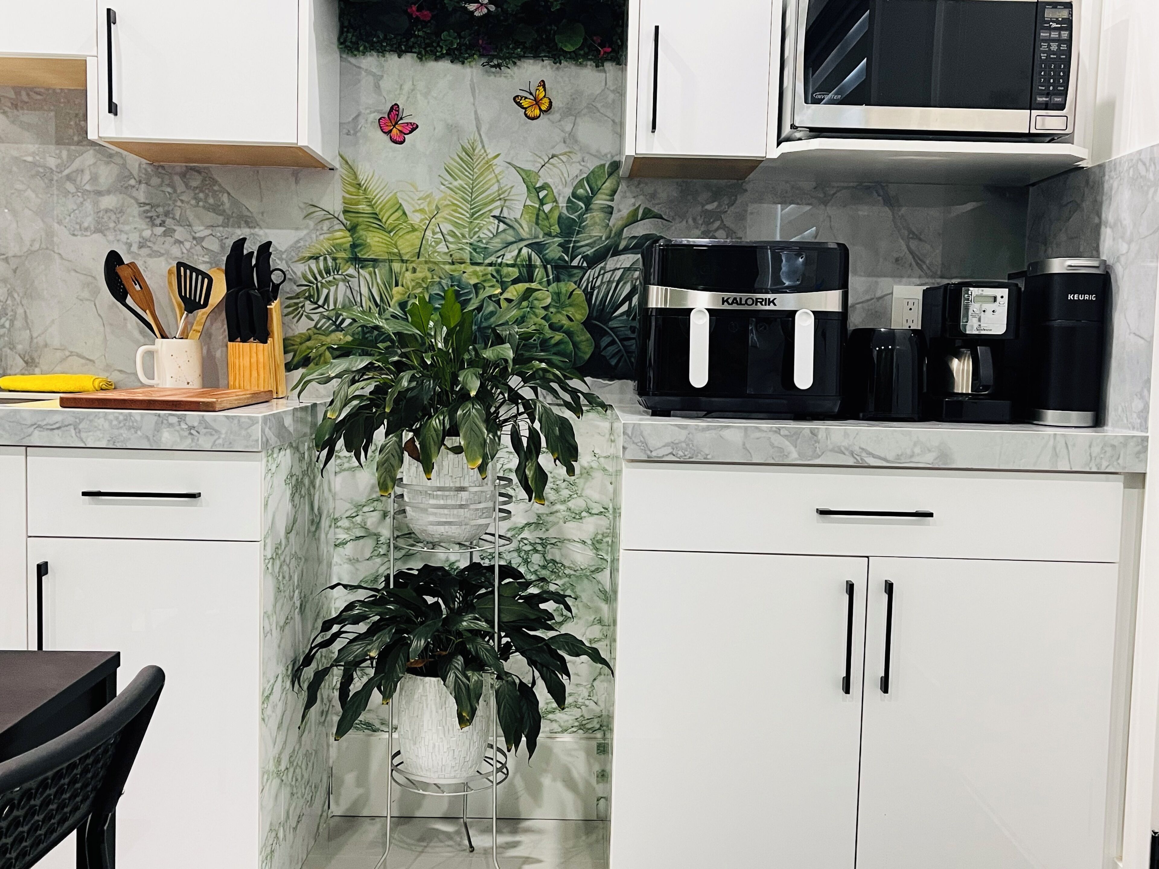 Une cuisine avec des meubles blancs, un four à micro-ondes, une machine à café et une plante en pot.