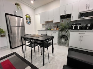 A modern kitchen with white cabinets, a stainless steel refrigerator, a dining table with chairs, and a coffee machine. - Peaceful 1BR Suite | Free Parking & Fast Wi-Fi (Delta)