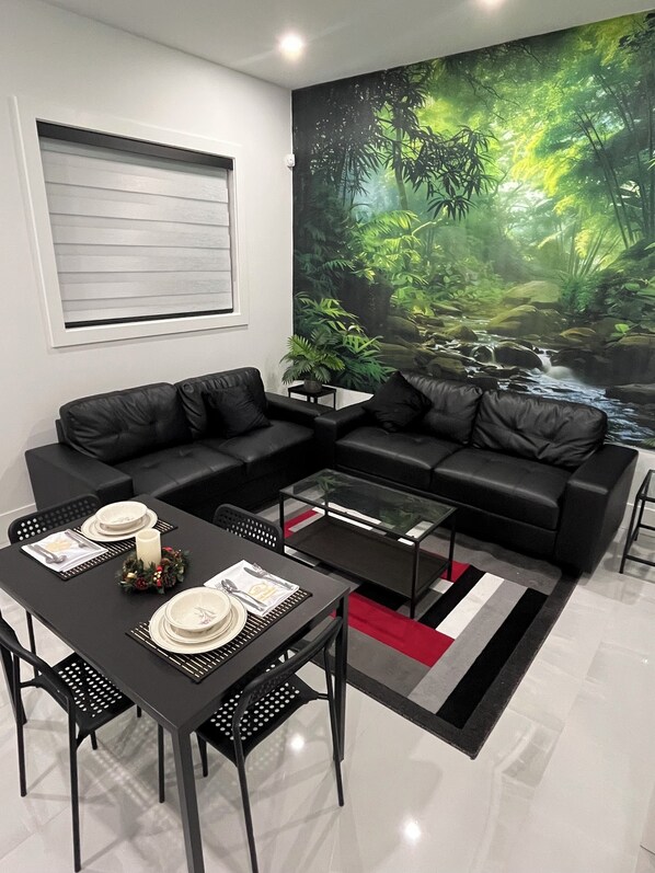 A modern living room with a black leather sectional sofa, a glass coffee table, and a dining table set for two. A large wall mural depicts a lush forest scene.