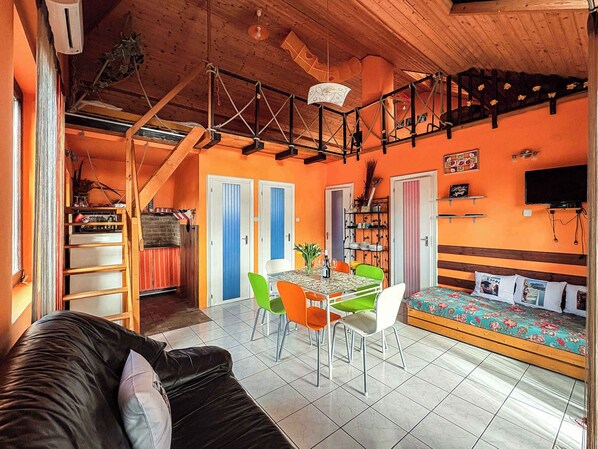 A cozy room with a wooden ceiling, a sofa, a dining table with chairs, and a bed with a floral bedspread.