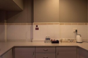 Uma cozinha de hotel com paredes revestidas de azulejos brancos, bancada com garrafas de água, chaleira e um escorredor de louça pequeno.