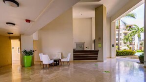 Spaziosa hall con reception, due poltrone bianche, un vaso con piante e vista su palme e edifici.