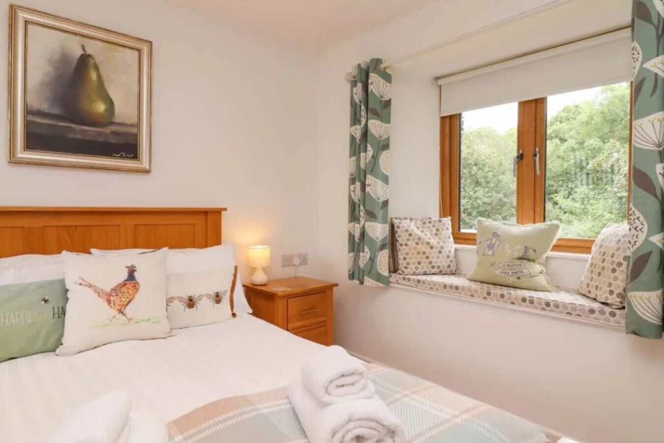 Un dormitorio con cama, almohadas, una mesita de noche, una lámpara, un rincón con cojines junto a la ventana y un cuadro en la pared.