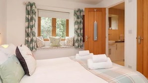A bedroom with a large bed, a window with curtains, a bedside table, and a bathroom visible through an open door. - Cawsand Cottage in Cornwall (Saltash)