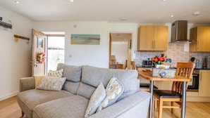 A modern living room with a grey sofa, wooden dining table, and a kitchen area with wooden cabinets and a stainless steel range hood. - Cawsand Cottage in Cornwall (Saltash)