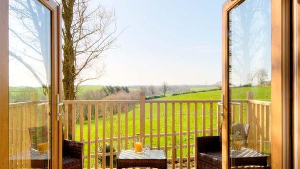 A balcony with a view of a green field, a wooden fence, and two wicker chairs with a table. - Cawsand Cottage in Cornwall (Saltash)