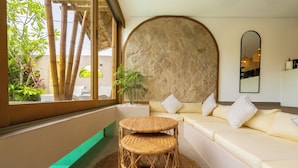 A room with a large window, a round wicker table, a sofa with cushions, and a stone wall. - Sunset Bamboo by Pertama Stay (Kerobokan)