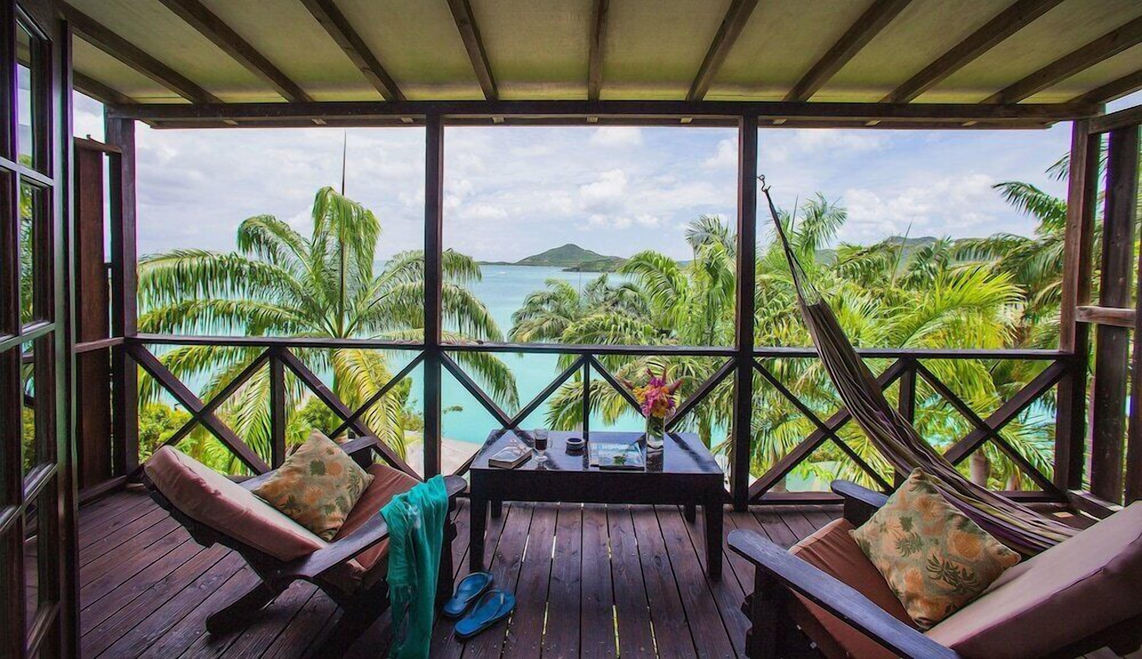 A wooden deck with two cushioned chairs and a table, overlooking a tropical beach with palm trees and a clear view of the ocean.