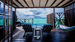 Eine Holzterrasse mit zwei Stühlen und einem Tisch, mit Blick auf das türkisblaue Meer unter einem bewölkten Himmel.