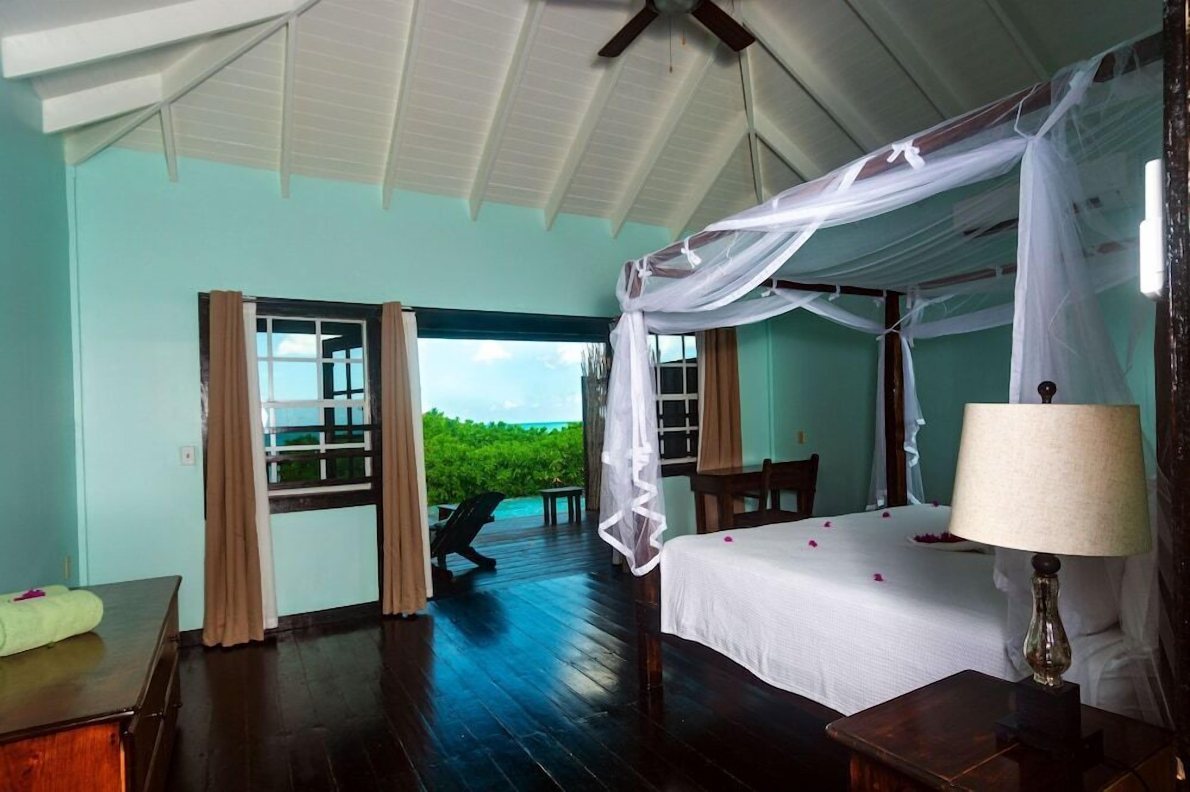 A four-poster bed with a canopy, a bedside table with a lamp, and a view of the ocean through a window.