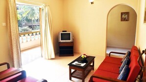 A living room with a sofa, a coffee table, a TV on a stand, and a balcony with curtains. - Lillywoods Highland Beach Resort (Candolim)