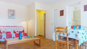 Una sala acogedora con un sofá, una mesa de centro de madera y una mesa de comedor con sillas. Hay una puerta que da a otra habitación y una lámpara en el suelo.