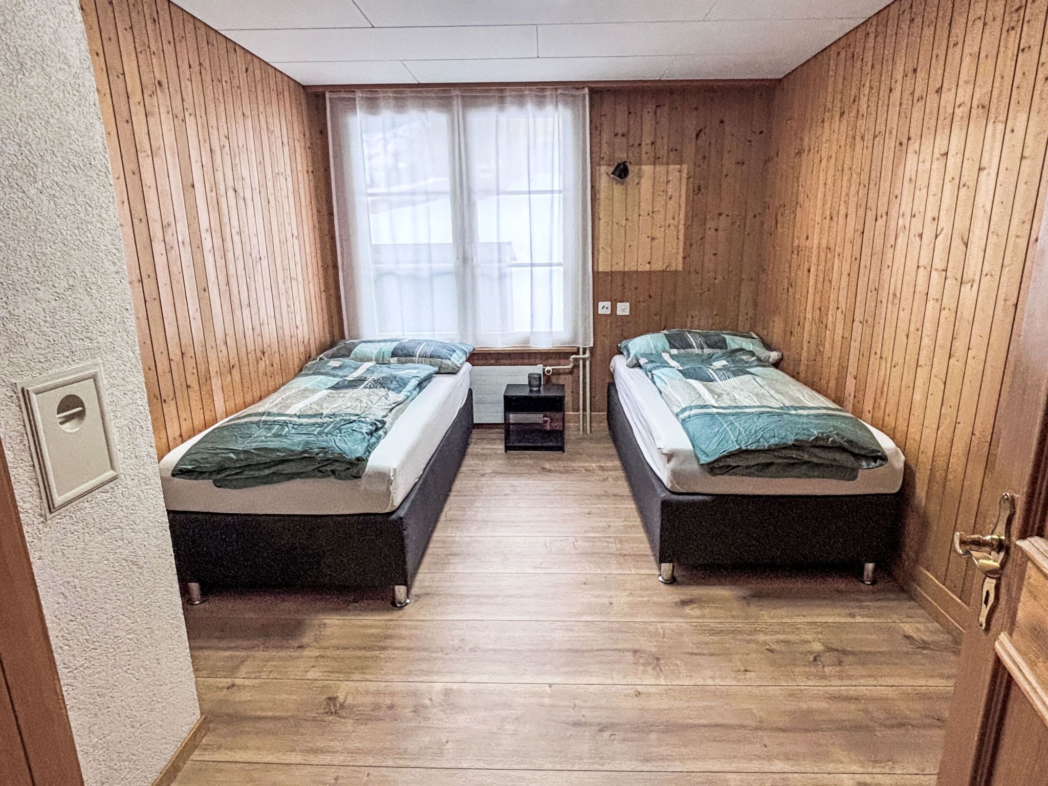 A small, compact room with two single beds, a small table, and a window with curtains.