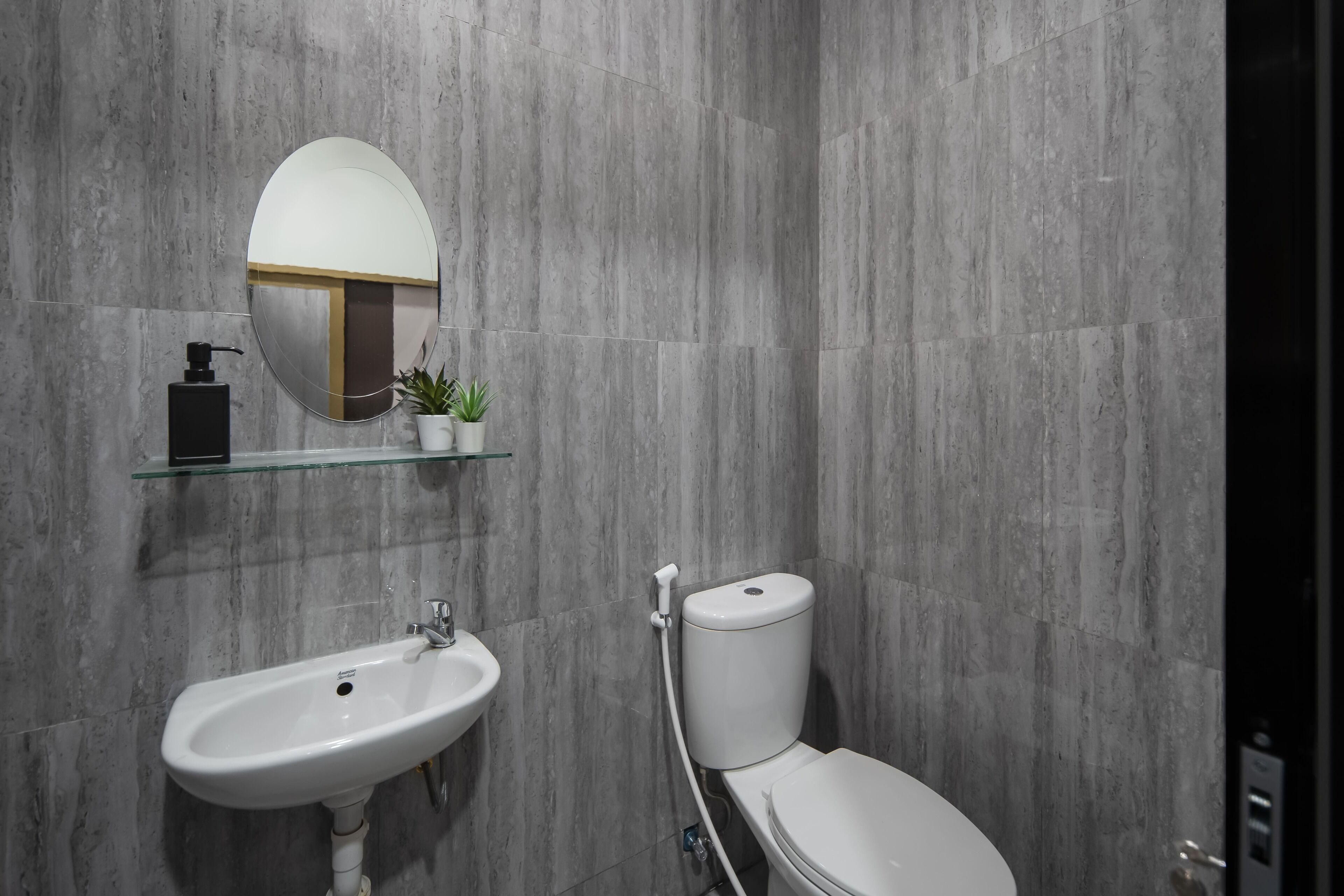 A modern bathroom with a white sink, a toilet, a round mirror, and a glass shelf with a small plant.