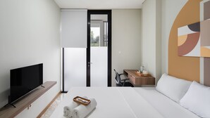 A modern hotel room with a flat-screen TV, a neatly made bed, a desk with a chair, and a view through an open door.
