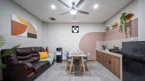 A modern living room with a sofa, coffee table, and dining table. There is a kitchen area with a refrigerator, stove, and shelves.