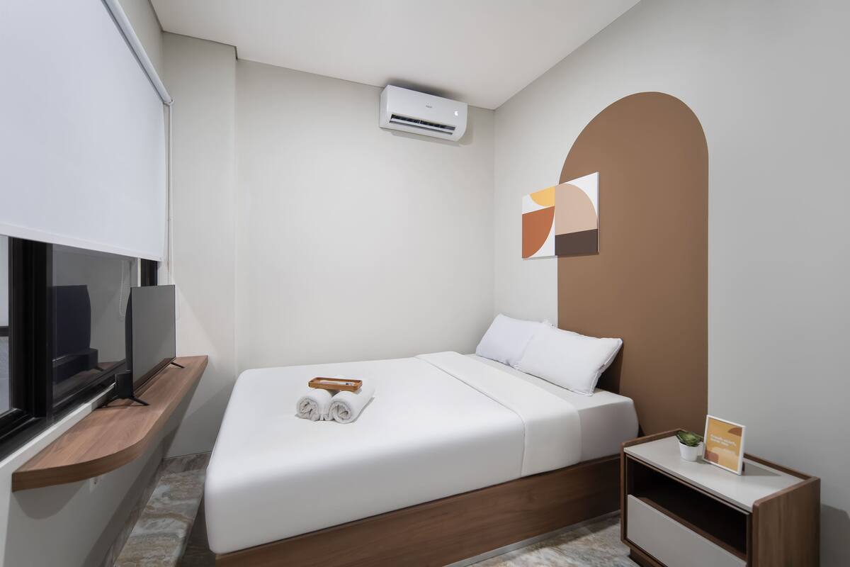 Moderni hotellihuone, jossa on sänky, puinen hylly, jonka päällä on televisio, ilmastointilaite ja pieni pöytä, jonka päällä on kasvi.