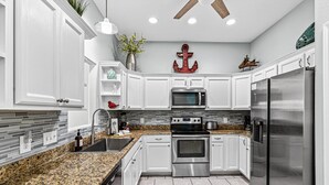 Uma cozinha moderna com armários brancos, bancadas de granito, eletrodomésticos de aço inoxidável e um detalhe decorativo de âncora na parede.