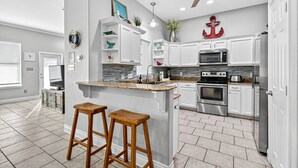 Uma cozinha moderna com armários brancos, bancada de granito e eletrodomésticos de aço inoxidável. Há dois banquinhos de madeira e um ventilador de teto.