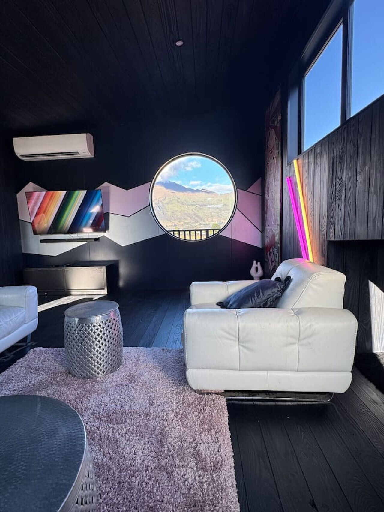 A modern living room with a round window, white leather sofa, and a view of a mountainous landscape.