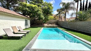 Una piscina rectangular con borde de baldosas, rodeada de césped artificial, y un edificio blanco con una ventana y una puerta.