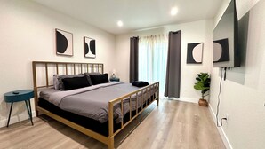 Un dormitorio moderno con estructura de cama de madera, una mesita de noche, televisor montado en la pared y una planta en maceta.