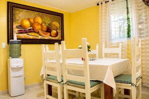 A dining area with a table, chairs, a water dispenser, and a fruit painting on the wall. - Urban Villa with Private Hot Tub & 6 Bedrooms (Santo Domingo)