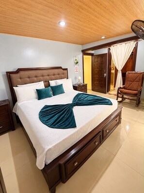 A neatly made bed with a wooden headboard, a nightstand with a lamp, and a chair in a well-lit room with a wooden ceiling and a door leading to another room.