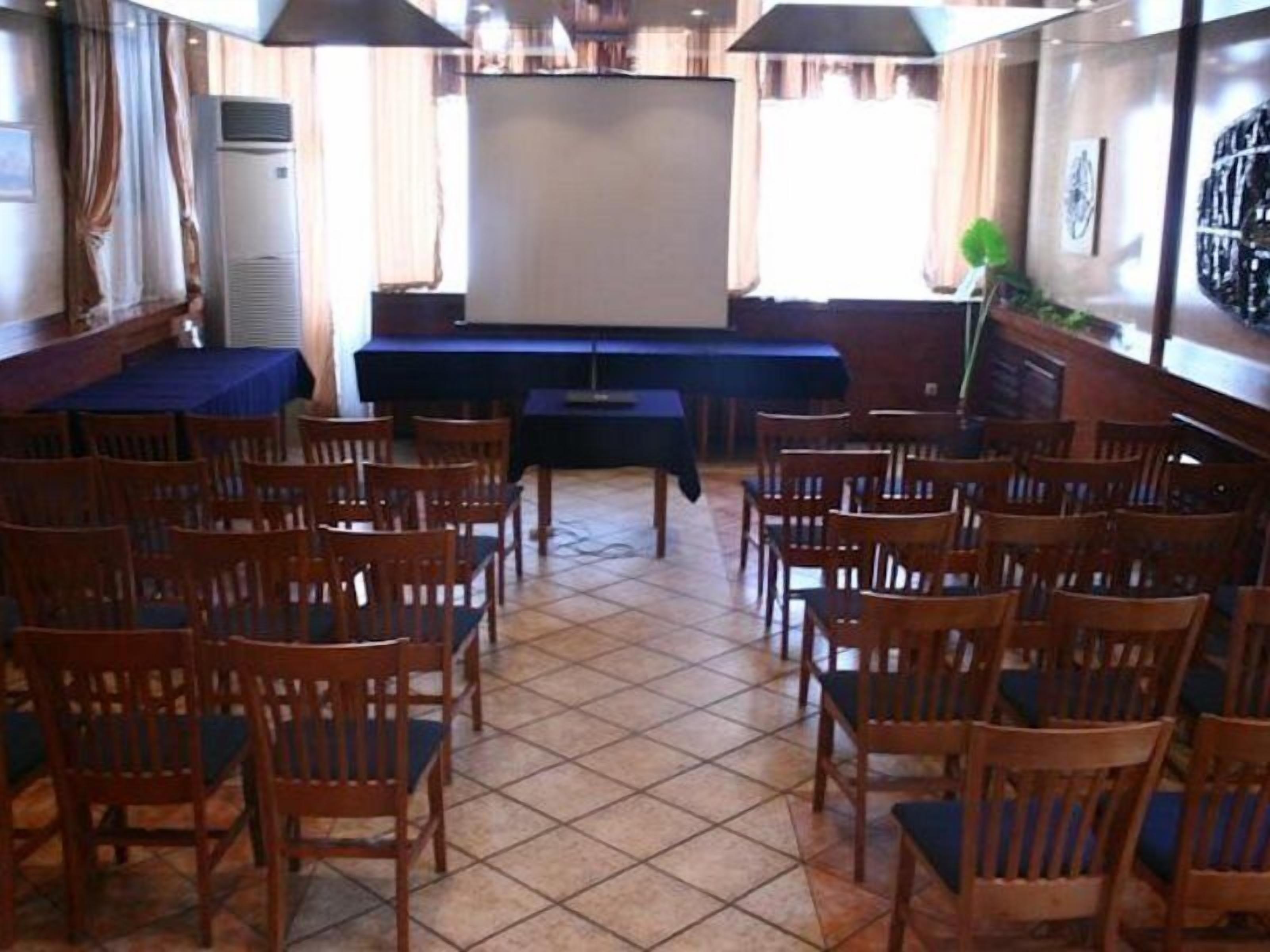 A room with wooden chairs arranged in rows, a projector screen, and a table with a blue tablecloth.