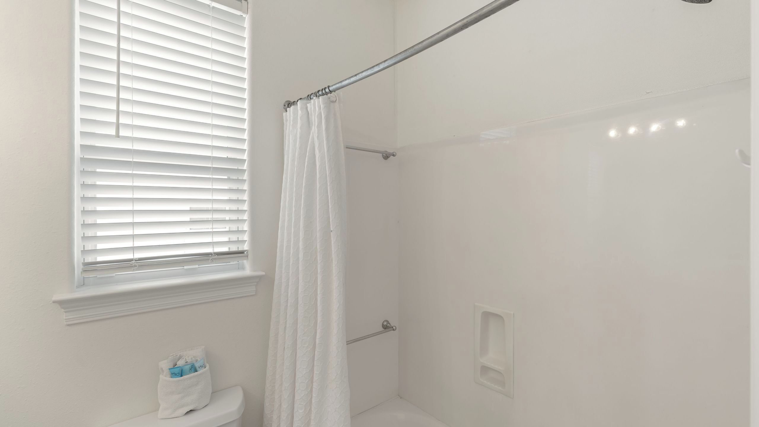 A bathroom with a shower, toilet, and window with blinds.