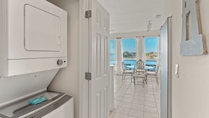 Una habitación con lavadora, una pequeña mesa con una tarjeta, una puerta que da a un balcón con vistas al agua y edificios.