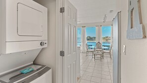 A room with a washing machine, a small table with a card, a door leading to a balcony with a view of a body of water and buildings. - Emerald Villa by Avantstay Lakefront + Communal Pool + Balcony Views + Near the Beach (Destin)