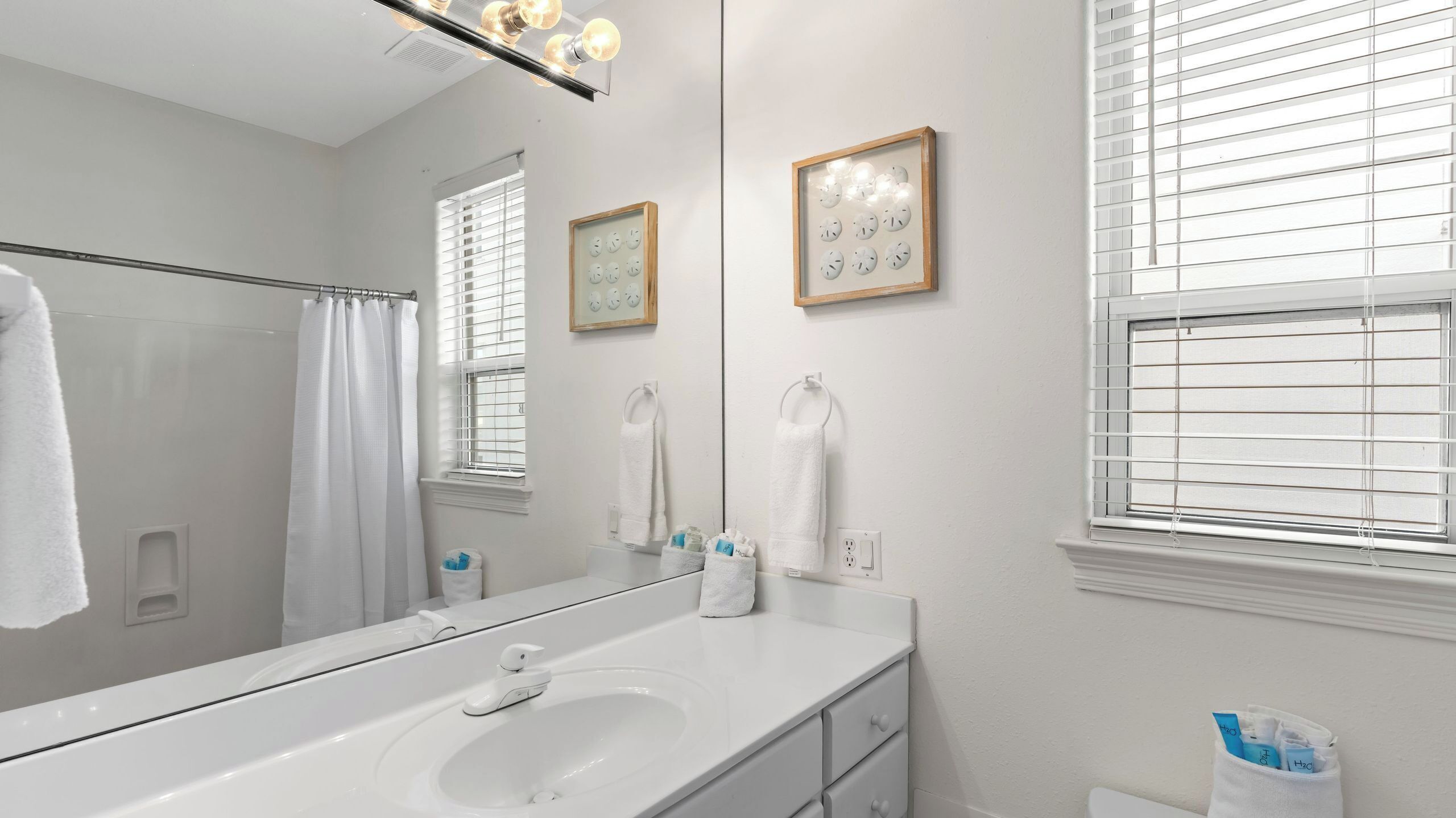 A modern bathroom with a large mirror, white fixtures, and a window with blinds.