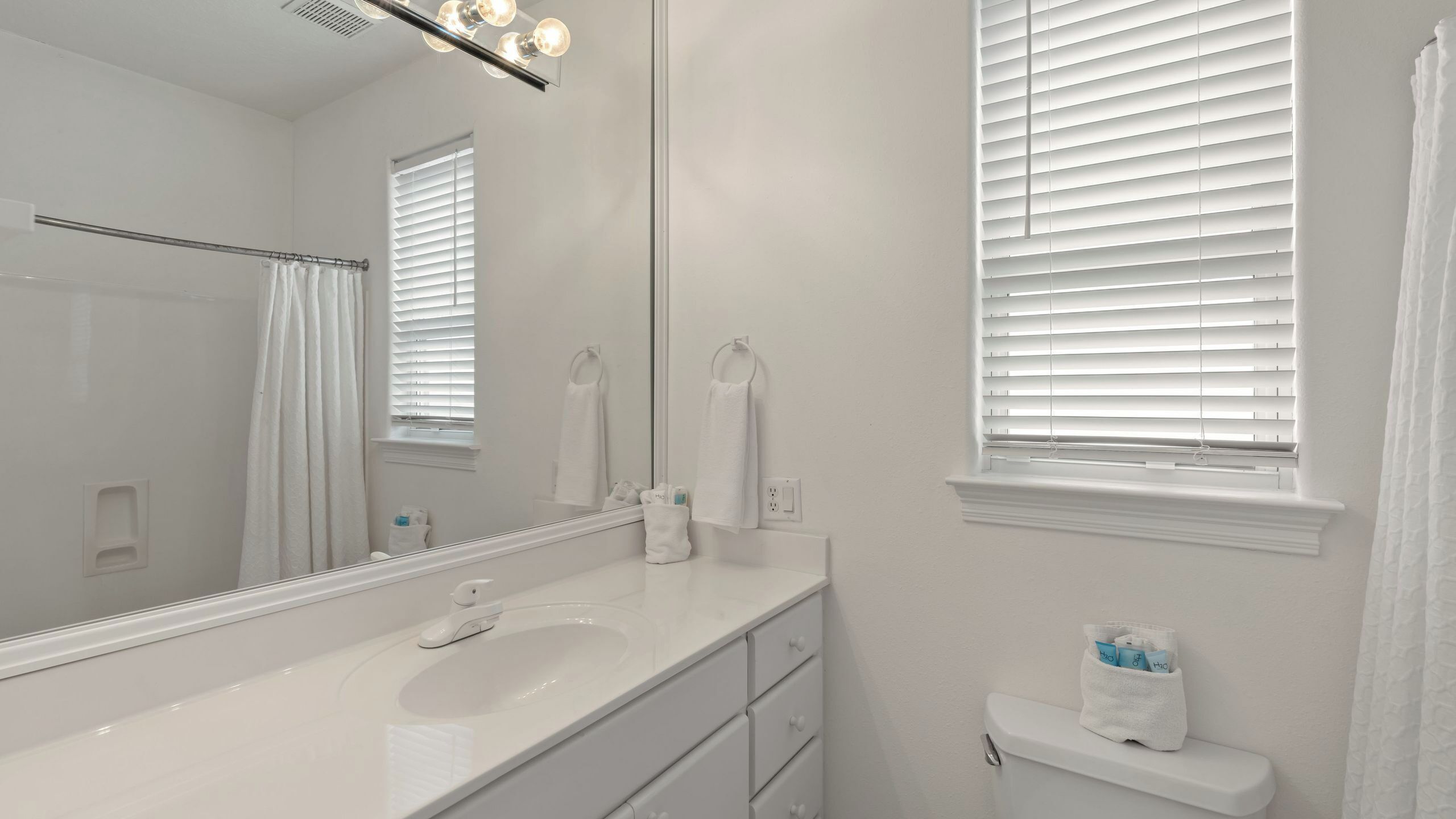 Un baño con lavamanos blanco, espejo y ventana con persianas.