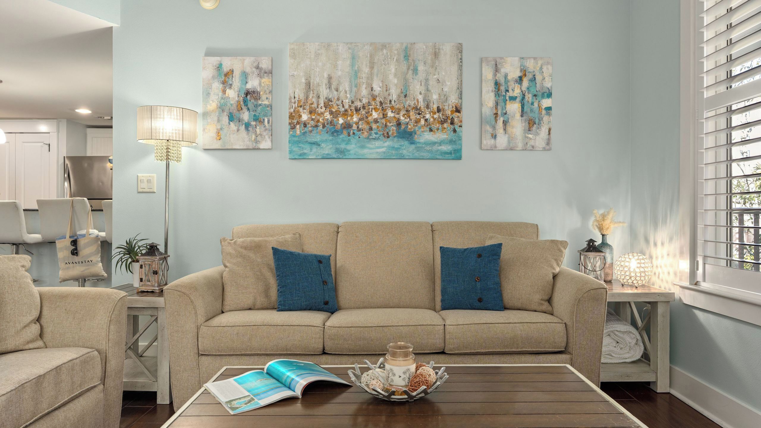 A living room with a beige sofa, two blue cushions, a coffee table with a magazine, and abstract wall art.